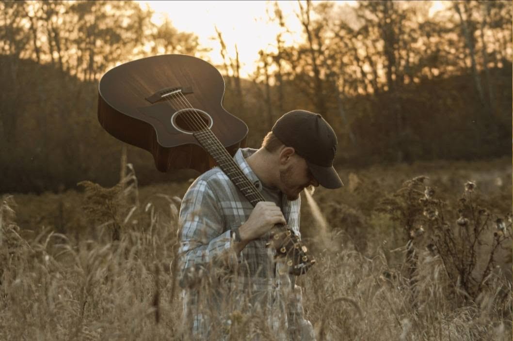 Robbie Jordan carrying an acoustic guitar through a sunlit field